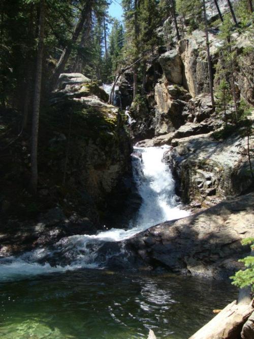 A serene waterfall cascading over rocks into a clear pool, surrounded by lush green trees and rocky terrain.