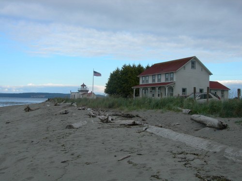 A sandy beach with a lighthouse and a house nearby, surrounded by grass and trees under a cloudy sky.