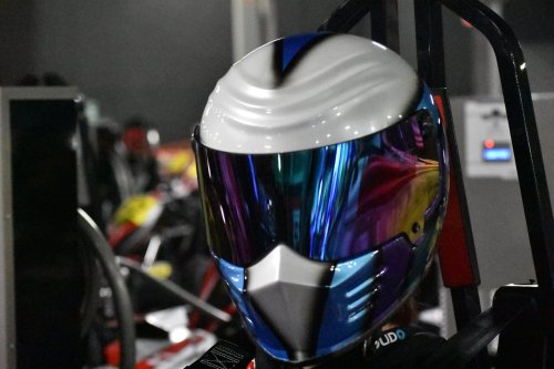 A close-up of a colorful racing helmet with a reflective visor, set against a blurred background of racing karts.
