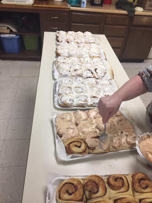 A table with several trays of cinnamon rolls, some being frosted, in a kitchen setting.