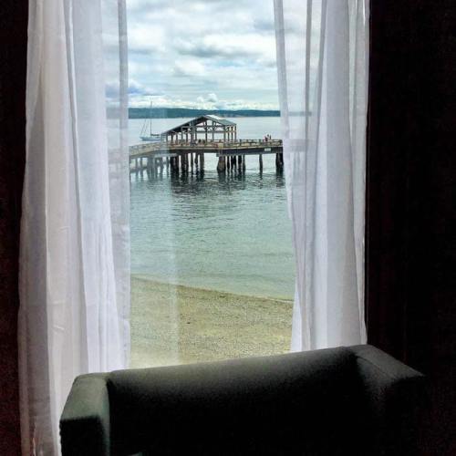View of a serene beach and pier through sheer white curtains, with a calm sea and cloudy sky in the background.
