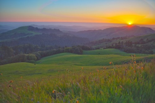 A vibrant sunset over rolling green hills, with a colorful sky and distant mountains in the background.