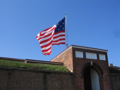 A large American flag waves atop a historic brick fort against a clear blue sky.