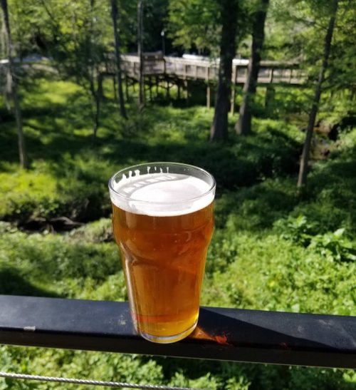 A glass of amber beer sits on a railing, with a lush green landscape and trees in the background.