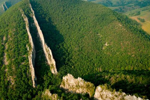 Aerial view of a lush green landscape featuring steep, rocky cliffs and a narrow, vertical rock formation.