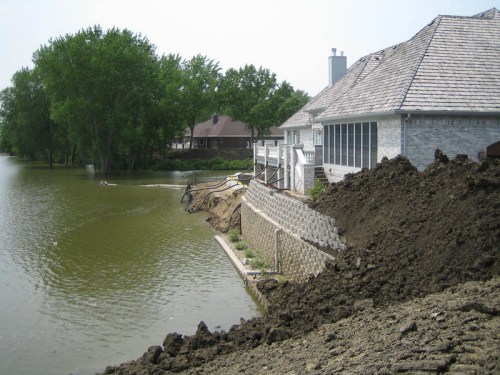 A house by a lake with a steep, muddy bank and trees in the background, showing signs of erosion and high water levels.