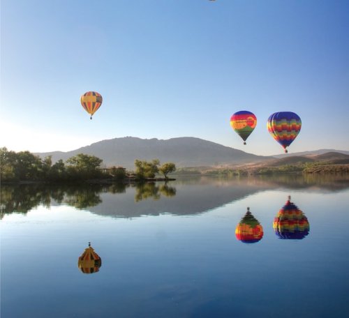 Colorful hot air balloons float above a calm lake, reflecting on the water with mountains in the background.