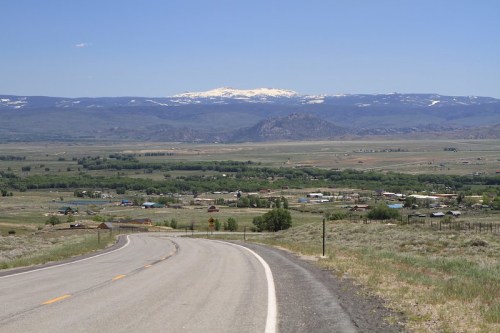 A winding road leads to a valley with small buildings, green fields, and snow-capped mountains in the background.