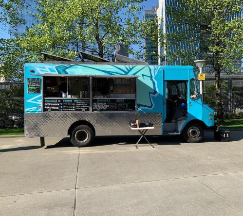 A vibrant blue food truck with a menu displayed, parked in a sunny area surrounded by trees and buildings.
