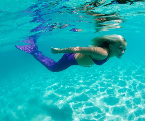 A young girl swims underwater, wearing a purple mermaid tail, with sunlight shimmering on the water's surface.