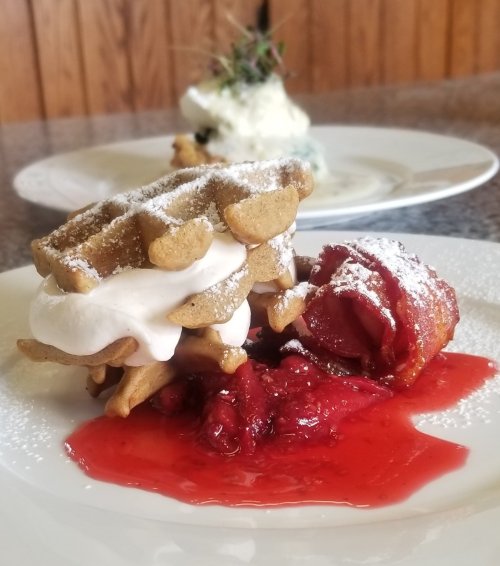 A plate featuring waffles with cream and strawberries, drizzled with red sauce, dusted with powdered sugar.