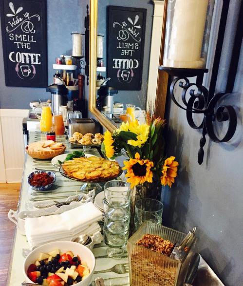 A buffet table with coffee, pastries, fruit, and flowers, set against a wall with decorative signs.