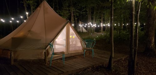 A cozy glamping tent at night, illuminated by string lights, with two turquoise chairs on a wooden deck.