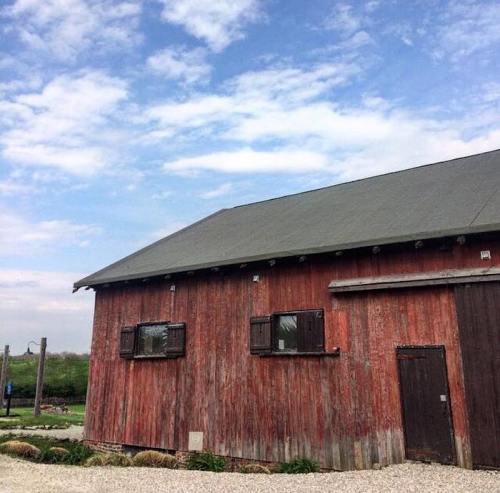 A rustic red barn with wooden shutters under a partly cloudy sky, surrounded by greenery and gravel.