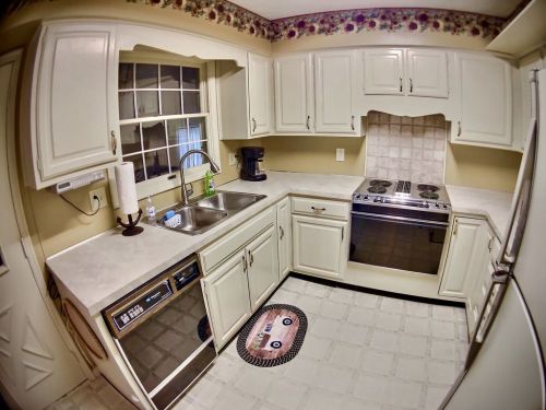 A cozy kitchen with white cabinets, a sink, stove, and a patterned rug on the floor. Natural light from a window.