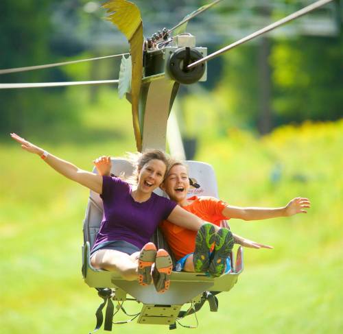 Two women joyfully riding a chairlift, arms outstretched, with green hills in the background.