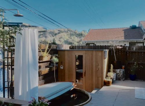 A serene backyard with a wooden sauna, outdoor shower, and a hot tub, surrounded by plants and mountains in the background.