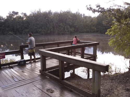 Two people fishing from a wooden dock by a calm, misty lake surrounded by trees.