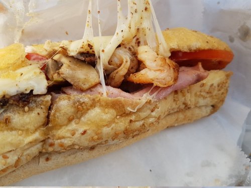 Close-up of a sandwich filled with shrimp, ham, melted cheese, and vegetables, served in a sesame seed bun.