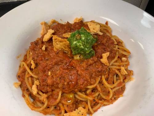 A plate of spaghetti topped with a rich tomato sauce, crispy chips, and a dollop of green pesto.
