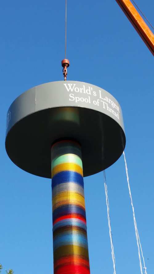 A colorful, oversized spool of thread is suspended from a crane against a clear blue sky.