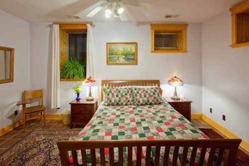Cozy bedroom with a quilted bedspread, two lamps, a chair, and a window, featuring warm lighting and decorative artwork.