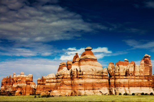 Rock formations with layered red and white cliffs under a blue sky with scattered clouds. Lush green grass in the foreground.