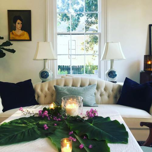 A cozy living room with a white sofa, decorative pillows, candles, and a large leaf centerpiece by a window.