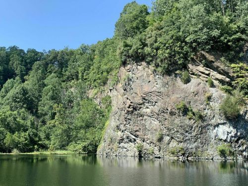 A serene lake surrounded by lush green trees and a rocky cliff under a clear blue sky.