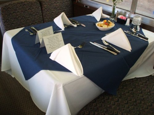 A neatly set dining table with a navy tablecloth, white napkins, silverware, and a bowl of fruit in the center.