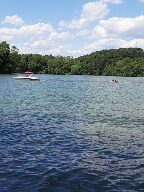 A boat and a person on a jet ski on a calm lake surrounded by green trees and a blue sky with fluffy clouds.