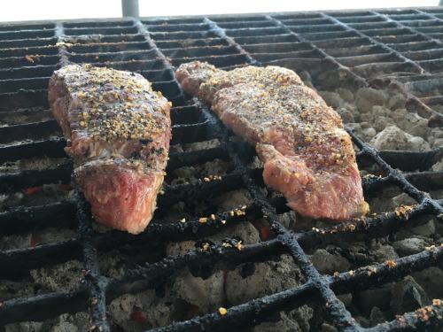 Two seasoned steaks grilling on a barbecue grate over hot coals, with a smoky background.