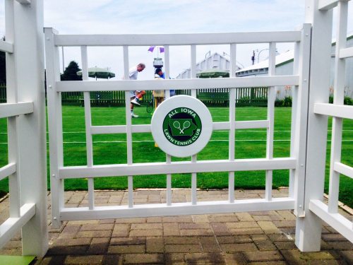 A white gate with a green sign reading "All Iowa Tennis Club" opens to a grassy tennis court in the background.