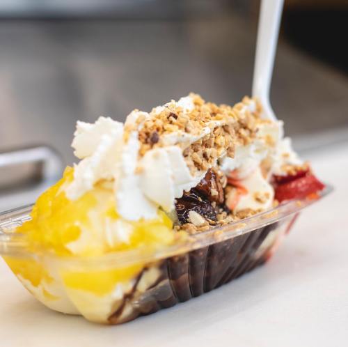 A colorful dessert with layers of ice cream, whipped cream, fruit, and granola in a clear cup.