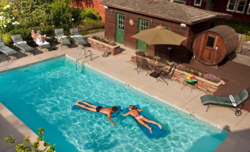Two people relax on pool floats in a clear blue pool, surrounded by lounge chairs and a wooden shed in a sunny backyard.
