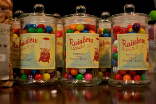 Colorful gumballs in glass jars labeled "Rainbow Gumballs" on a wooden surface.
