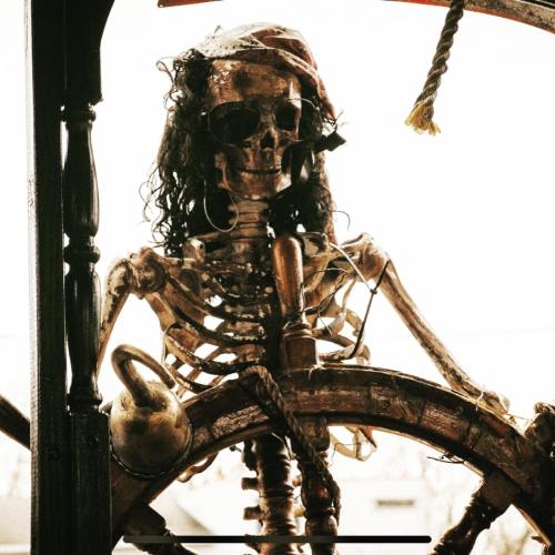 A skeleton dressed as a pirate stands at the helm of a ship, gripping the ship's wheel.