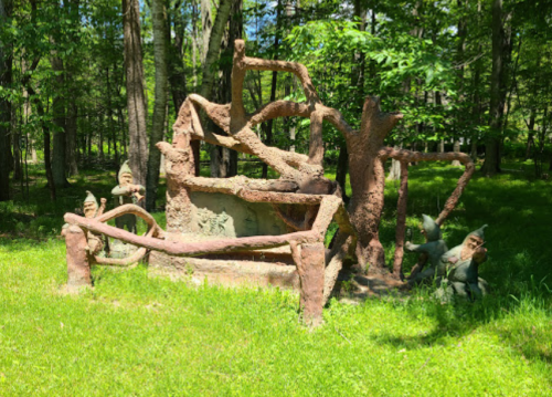 A whimsical sculpture in a forest, featuring tree-like structures and small figures among lush green grass.