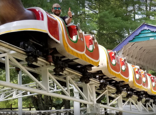 A person gives a thumbs up while riding a colorful roller coaster in a wooded amusement park setting.