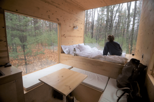 Cozy wooden cabin interior with a bed and large windows overlooking a forest, person gazing outside.