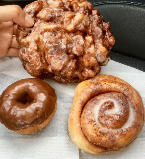 A hand holding a large apple fritter next to a chocolate donut and a cinnamon roll on a paper napkin.