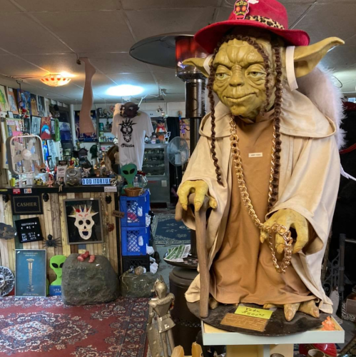 A life-sized Yoda statue in a hat and robe, surrounded by eclectic decor in a quirky shop.