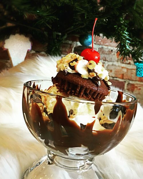 A glass dessert bowl filled with chocolate sauce, whipped cream, a cupcake, and topped with a cherry.