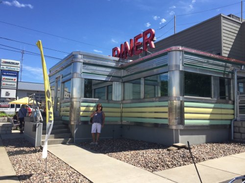 A woman stands outside a retro-style diner with a bright sign, featuring chrome and yellow accents on a sunny day.