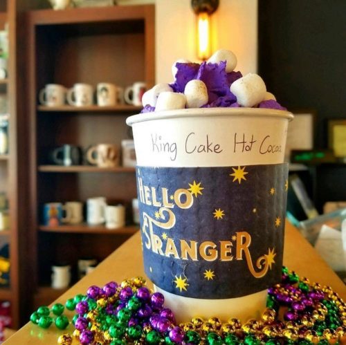 A cup of King Cake Hot Cocoa topped with purple whipped cream and mini marshmallows, surrounded by colorful beads.