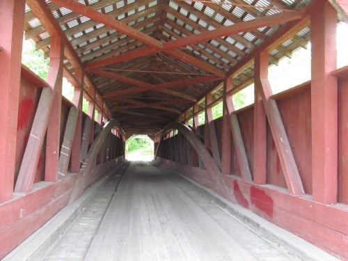 Interior view of a covered bridge with wooden beams and a dirt floor, leading to a green landscape outside.