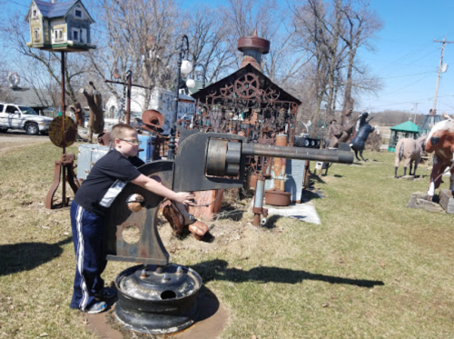 A child interacts with a large, vintage machine in an outdoor display area with sculptures and structures in the background.