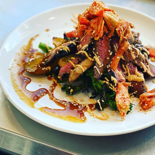 A gourmet dish featuring sliced beef, kimchi, sesame seeds, and drizzled sauce on a bed of greens and potatoes.