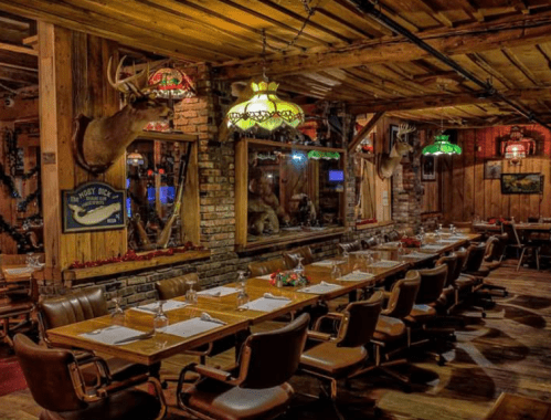 Cozy rustic restaurant interior with wooden decor, a long table set for dining, and mounted animal heads on the walls.