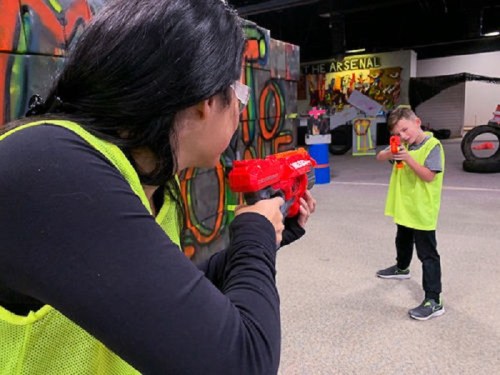 A woman and a boy aim toy guns at each other in a colorful indoor play area, both wearing bright green vests.